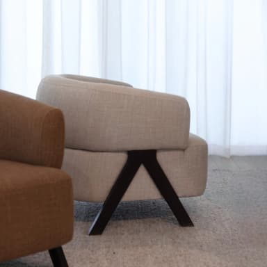 A modern armchair with a rounded, upholstered body and angled legs. The armchair features a curved backrest that flows into armrests, all covered in a textured light beige fabric. The base consists of two dark, V-shaped legs that are angled outwards, providing a stable support. The armchair is placed on a textured grey carpet. — 3D model
