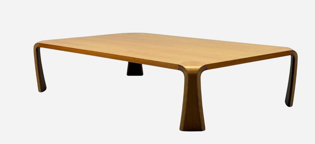 A modern rectangular coffee table constructed from molded plywood with a light wood veneer finish. It features a seamless design where the tabletop curves down to form the legs, complemented by two discrete dark central supports. — 3D model