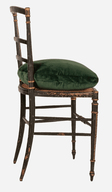 An antique Regency style side chair featuring an ebonized (black lacquered) wood frame with extensive gilt decoration. The chair has a slat back design with three horizontal slats adorned with geometric gilt patterns, complemented by reeded and gilt uprights and finials. The seat appears to be caned, currently topped with a loose, plush dark green velvet cushion. The front legs are turned with gilt accents, while the splayed rear legs are plain. Stretchers connect the legs, some with ring-turned details, and the ebonized finish shows visible distressing and wear. — 3D model