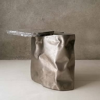A unique accent table featuring a heavily textured, crumpled cast metal base and an irregularly shaped, thick textured glass top. The base has an organic, fluid form with intentional creases and folds, finished in a dark, metallic tone. The glass top is clear, displaying a molten or rippled surface texture, and extends assymetrically over one side of the base, creating a dynamic visual balance. The design is contemporary and sculptural, making it a focal point. — 3D model