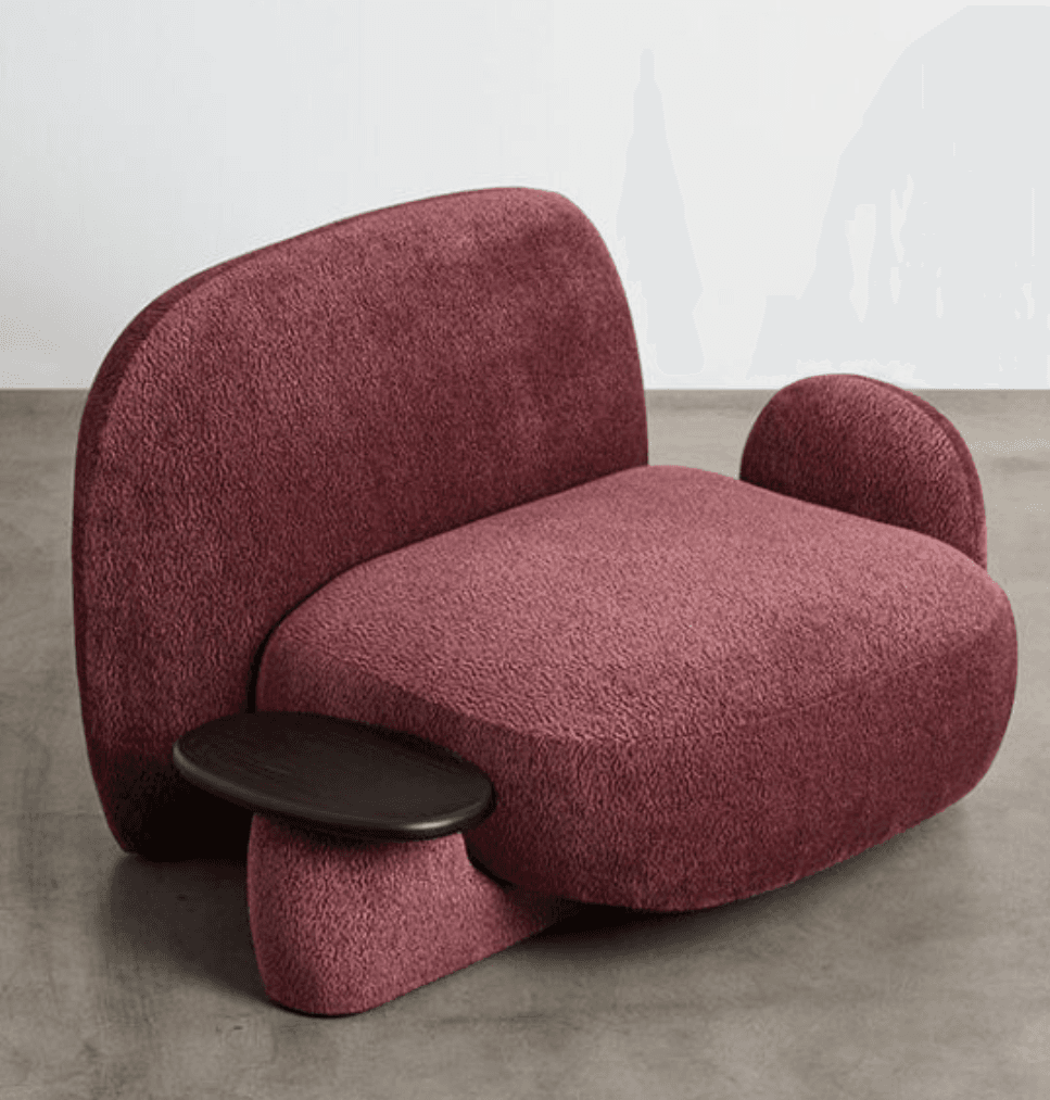 A contemporary armchair with a distinctive, organic silhouette. It features plush, textured upholstery in a deep rose or burgundy hue. The chair has a high, curved backrest and a rounded seat and armrests. A notable feature is the integrated, dark-finished oval side table attached to the left side of the base. — 3D model