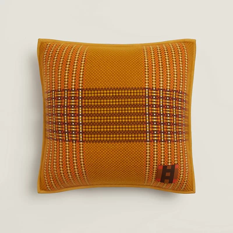 A square decorative cushion featuring a prominent plaid pattern in shades of mustard yellow, burnt orange, deep brown, and off-white. The cushion has a tightly woven texture and is finished with solid mustard yellow piping around its edges. A small, square brand tag with a stylized 'H' is visible in one corner. — 3D model