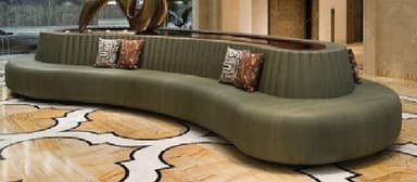 A contemporary, curved modular sofa with an integrated table running along its backrest. The seating is upholstered in a smooth olive green fabric, featuring a ribbed design on the back support where it meets the table. The integrated table has a dark brown, possibly wooden or laminate, surface. Several small decorative cushions with abstract patterns are placed on the seating. — 3D model