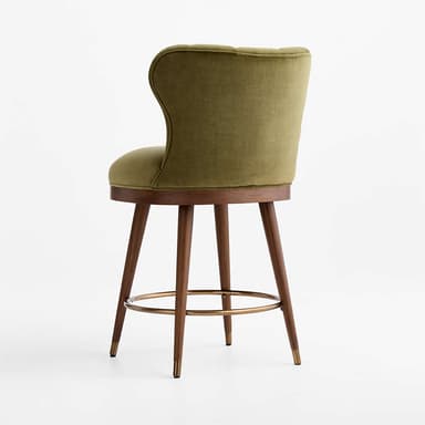 A stylish bar stool featuring an olive green velvet upholstered seat and a distinctive scalloped, channel-tufted backrest. It stands on four angled wooden legs, connected by a circular brass footrest, and finished with brass ferrules on the feet. The design blends luxurious materials with mid-century modern influences. — 3D model