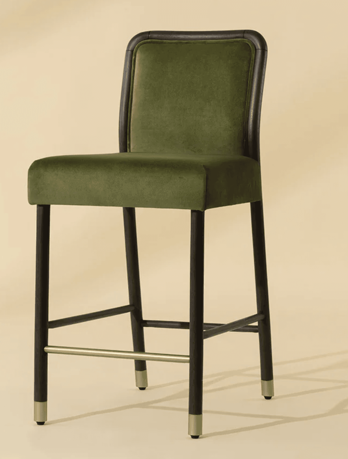 A contemporary bar stool featuring olive green velvet upholstery on both the seat and a rounded backrest. The frame is constructed from dark-stained wood, possibly black oak, with a distinctive curved detail around the backrest. The four tapered legs are also dark wood and are reinforced by stretchers, including a prominent brass-finished footrest. Each leg is capped with a subtle brass ferrule. — 3D model