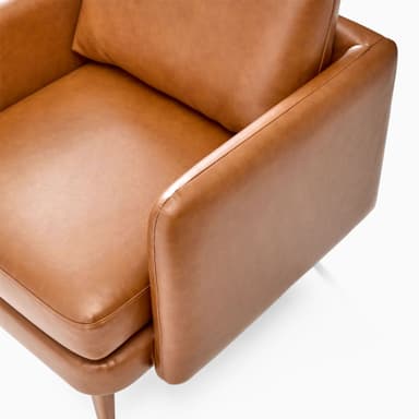 A modern armchair upholstered in brown leather. It features a boxy frame with rounded armrests and a slightly reclined backrest. The seat is composed of two thick cushions. The armchair is supported by four tapered wooden legs, angled outwards. — 3D model
