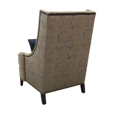 A fully upholstered wingback armchair, featuring a high back with subtly flared wings and rolled arms. It is covered in a textured, greenish-beige fabric, likely velvet or chenille. Decorative nailhead trim runs along the armrests and the bottom edge of the chair's frame. The chair rests on short, dark, tapered wooden legs. It is adorned with two accent pillows: one large rectangular pillow with a grey and beige abstract pattern, and a smaller solid navy blue square pillow with a central button tuft. — 3D model