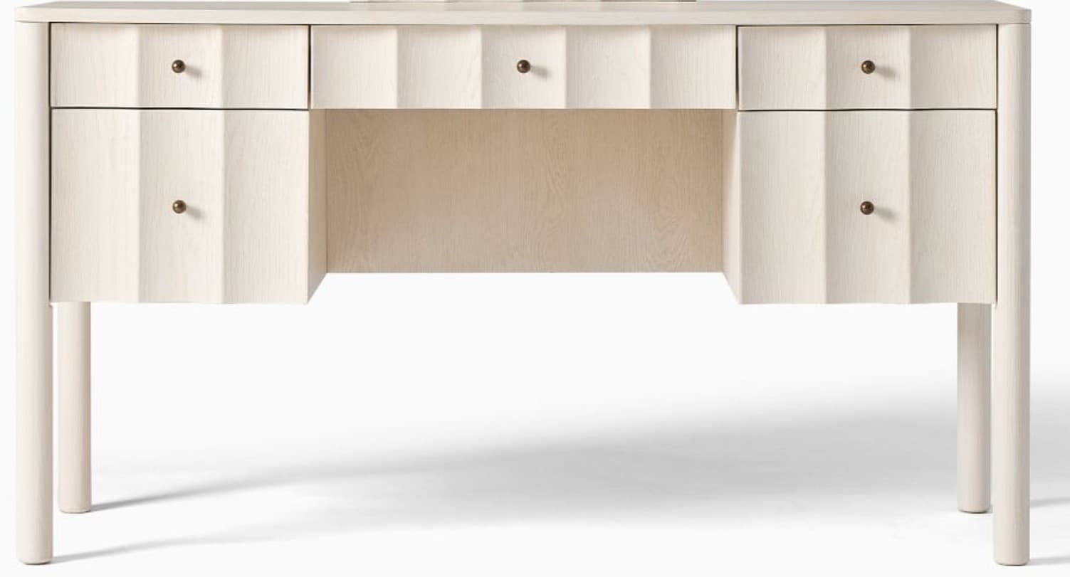 A light-colored, contemporary desk featuring a rectangular top with subtly rounded corners, supported by four tapered cylindrical legs. The desk includes multiple drawers, with the drawer fronts showcasing a distinct fluted or scalloped design, creating a textural element. Small, dark-toned spherical drawer pulls are present on each drawer. The overall aesthetic is clean and modern, with a focus on tactile surface detail. — 3D model