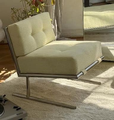 A modern armless lounge chair featuring a polished metal frame and thick, tufted cushions upholstered in ribbed, off-white fabric. The frame has a minimalist, open design with a sled-like base. The seat and back cushions are integrated into the frame, held by fabric straps on the lower front bar. — 3D model