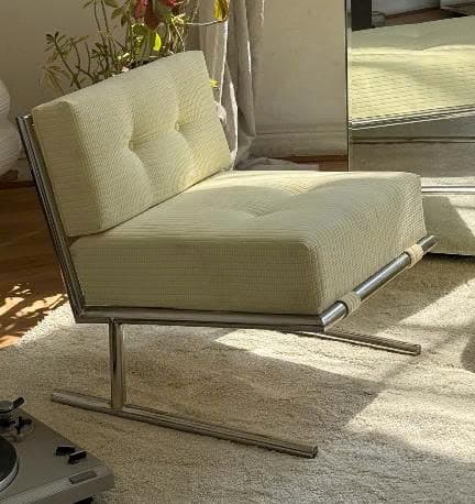 A modern armless lounge chair featuring a polished metal frame and thick, tufted cushions upholstered in ribbed, off-white fabric. The frame has a minimalist, open design with a sled-like base. The seat and back cushions are integrated into the frame, held by fabric straps on the lower front bar. — 3D model