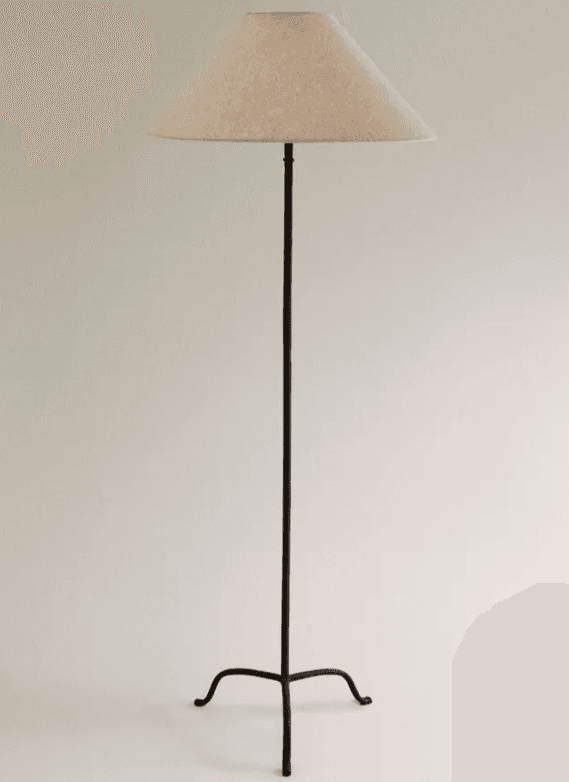 A tall, slender floor lamp featuring a dark, possibly wrought iron, stem that branches into a three-legged tripod base with gently curved feet. It is topped with a simple, conical lampshade made from a light-colored, textured fabric. — 3D model