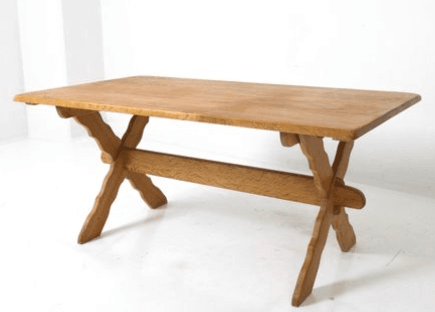 A rustic wooden dining table with a rectangular top and an X-shaped leg base. The legs are constructed from thick, joined wooden pieces, creating a stable and visually striking foundation. A stretcher bar connects the two X-leg assemblies, providing additional support and structural integrity. The wood exhibits a natural grain pattern and a warm, light brown finish. — 3D model