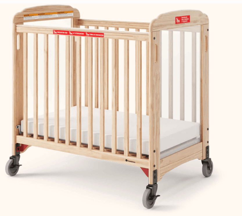 A compact, wooden crib with a white mattress. The crib features vertical slats along its sides and a slightly curved top rail. It is equipped with four wheels for mobility. The overall construction appears sturdy and functional, suitable for infants. — 3D model