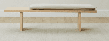 A minimalist wooden bench featuring a light-colored, rectangular cushion. The bench is constructed from light-toned wood with two solid plank legs supporting a long, flat top surface. — 3D model