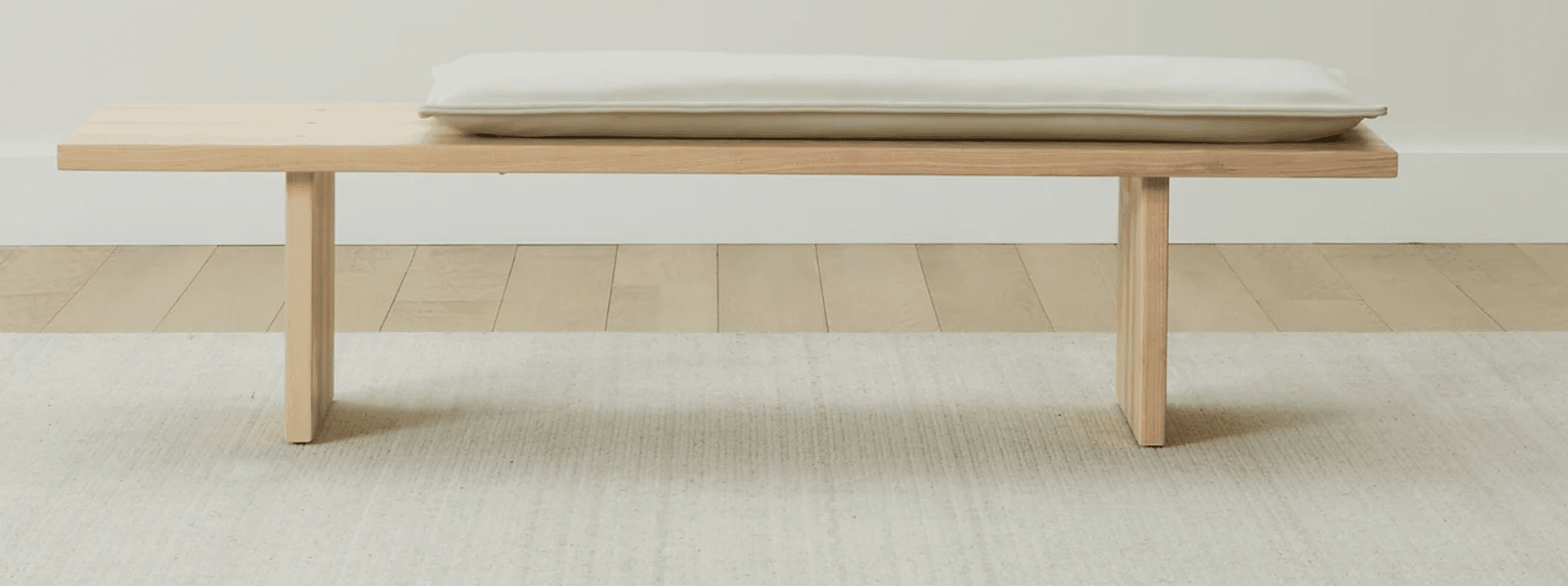 A minimalist wooden bench featuring a light-colored, rectangular cushion. The bench is constructed from light-toned wood with two solid plank legs supporting a long, flat top surface. — 3D model