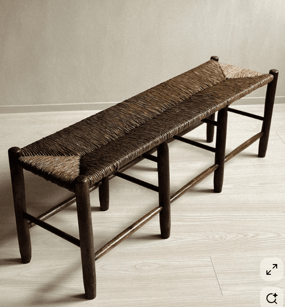 A long, narrow bench with a rush-woven seat. The frame and legs appear to be made of dark wood. The legs are cylindrical and taper slightly towards the floor. The seat is woven from natural fibers, likely rush or straw, in a pattern that creates texture and visual interest. The bench has a rustic and possibly traditional aesthetic. — 3D model