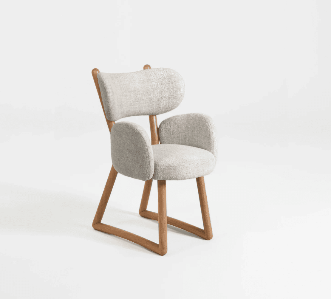 A modern armchair with a light grey upholstered seat, backrest, and armrests, supported by a natural wood frame. The frame has a distinctive angular design, particularly at the legs and where the backrest is supported. The upholstery has a textured fabric weave. — 3D model