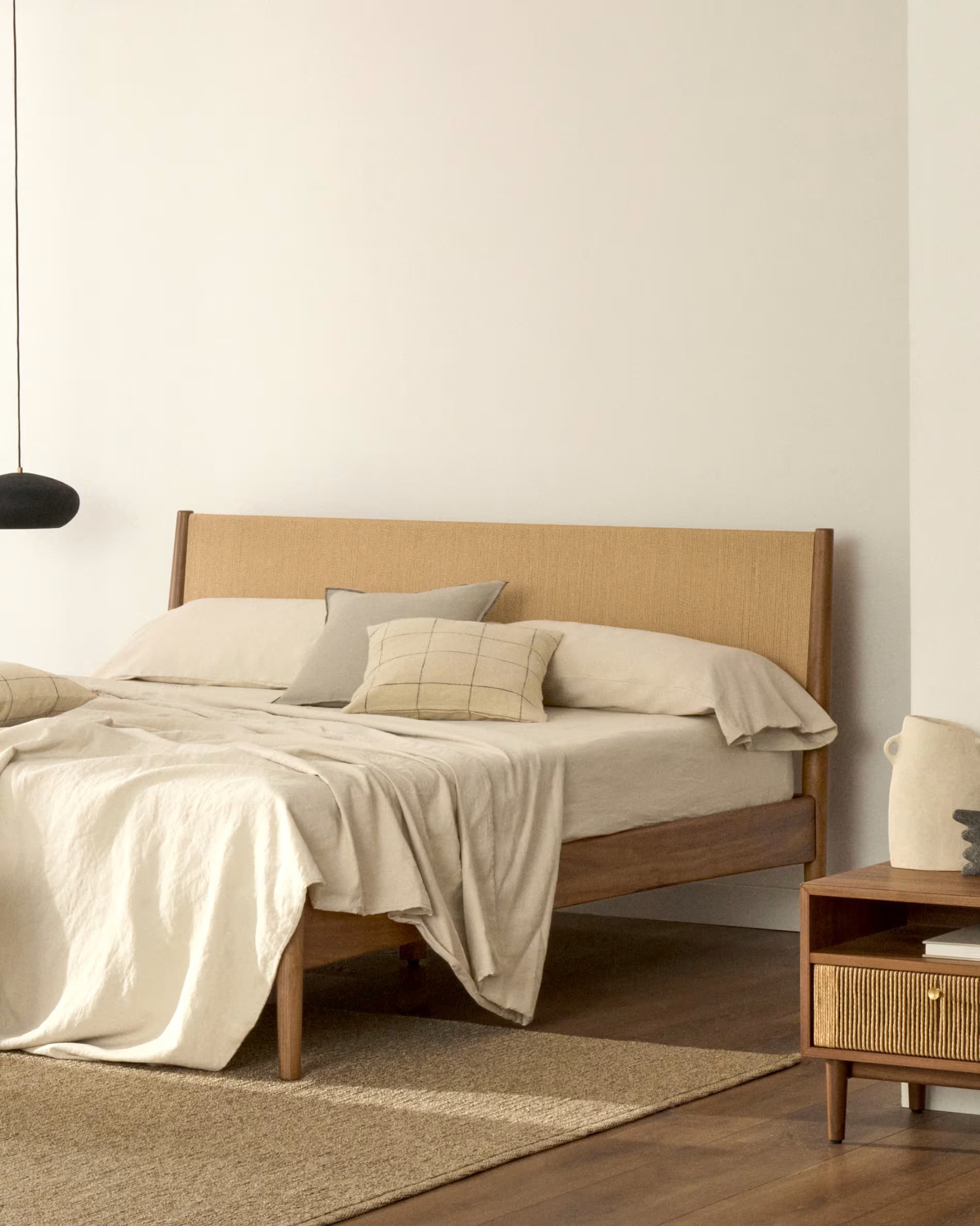 A modern platform bed featuring a clean-lined wooden frame and a headboard with a woven cane or rattan panel. The bed is presented with neutral-colored bedding, including a duvet, standard pillows, and two accent pillows. The frame has tapered legs and a low profile, contributing to a minimalist aesthetic. — 3D model