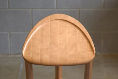 A wooden bar stool with a triangular, slightly dished seat and three cylindrical legs. One leg is positioned at the front and two at the back. A curved stretcher connects the two back legs near the bottom, and a diagonal stretcher connects the front leg to the rear left leg. The stool is constructed from light-colored wood with a visible grain. — 3D model