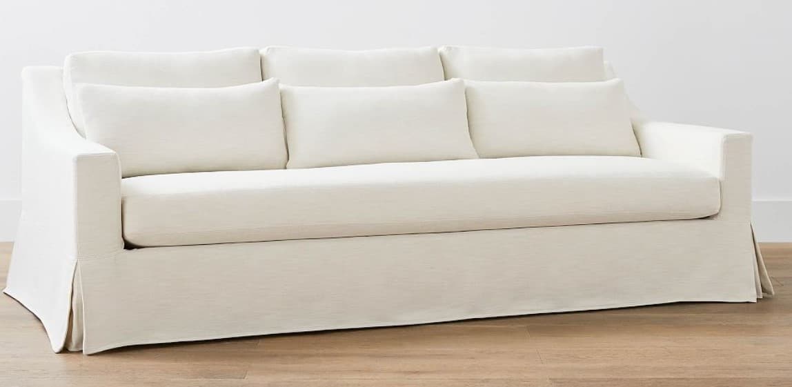 A three-seater sofa with a skirted base and loose back cushions. The sofa has track arms and is upholstered in a light-colored fabric. It is accompanied by three rectangular throw pillows. — 3D model