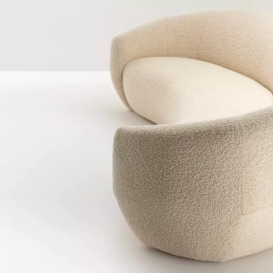 A cream-colored, curved sofa with a textured fabric upholstery. The sofa features a continuous, organic form with no visible legs, appearing to rest directly on the floor. Its design is modern and sculptural, with a rounded back and seat that create a sense of enclosure. — 3D model