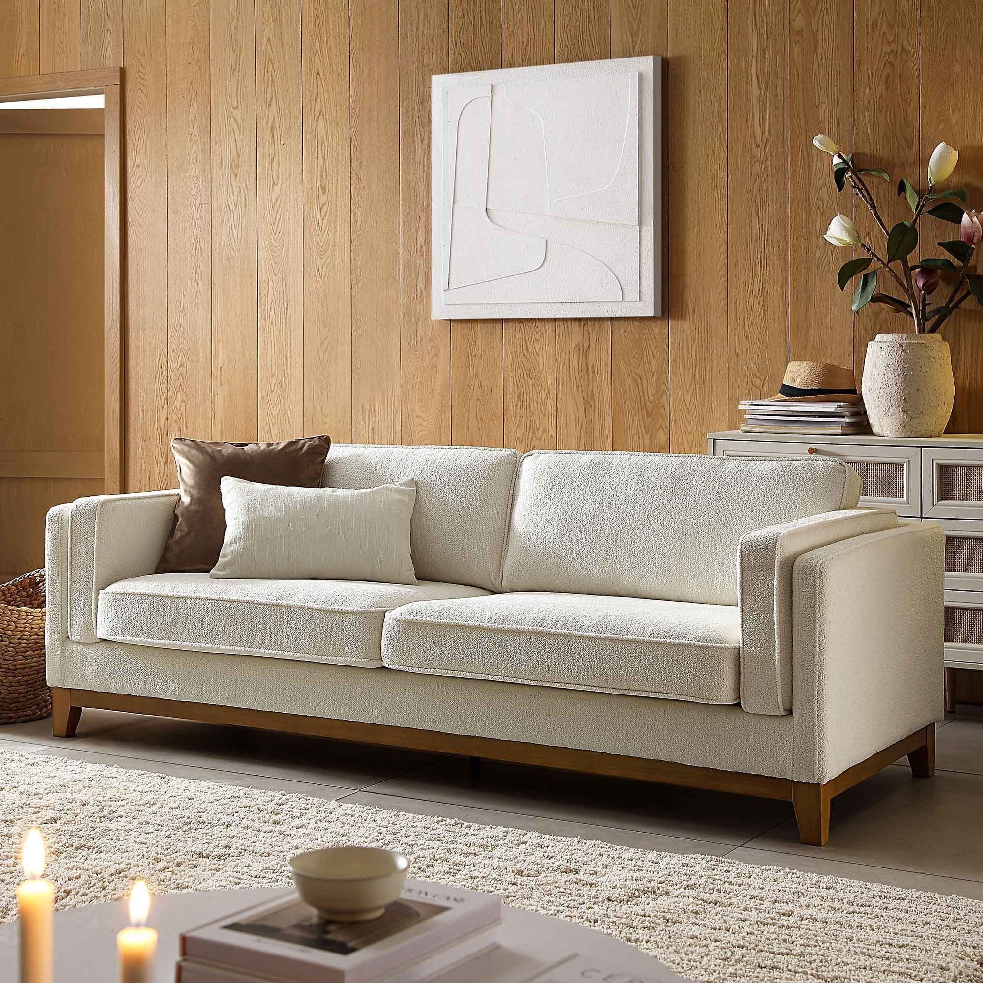 A modern three-seater sofa upholstered in a light cream-colored fabric with a subtle textured pattern. It features straight, clean lines with wide, rectangular arms and backrest, and two large seat cushions. The sofa rests on a recessed wooden base with four tapered wooden legs, providing a slight elevation. Two accent pillows, one in a solid cream and the other in a warm brown, are placed on the left side of the sofa. — 3D model