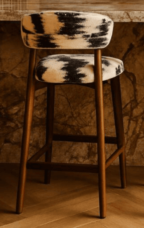 A modern bar stool featuring a sturdy wooden frame and distinctive cowhide-patterned upholstery on both the seat and the low, curved backrest. The stool has four tapered wooden legs connected by horizontal stretchers that double as footrests. — 3D model