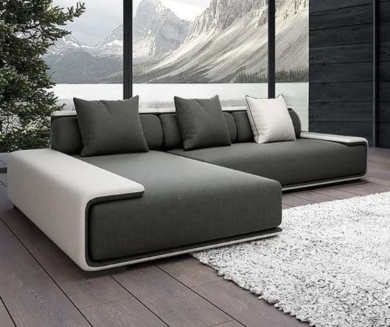 An L-shaped modern sectional sofa featuring dark gray fabric upholstery and a contrasting light-colored frame that forms integrated console arms and back support. The sofa has a low profile, comprising a main seating section and a chaise lounge component. It includes several square throw pillows, some matching the dark gray upholstery and one in white with dark piping. — 3D model