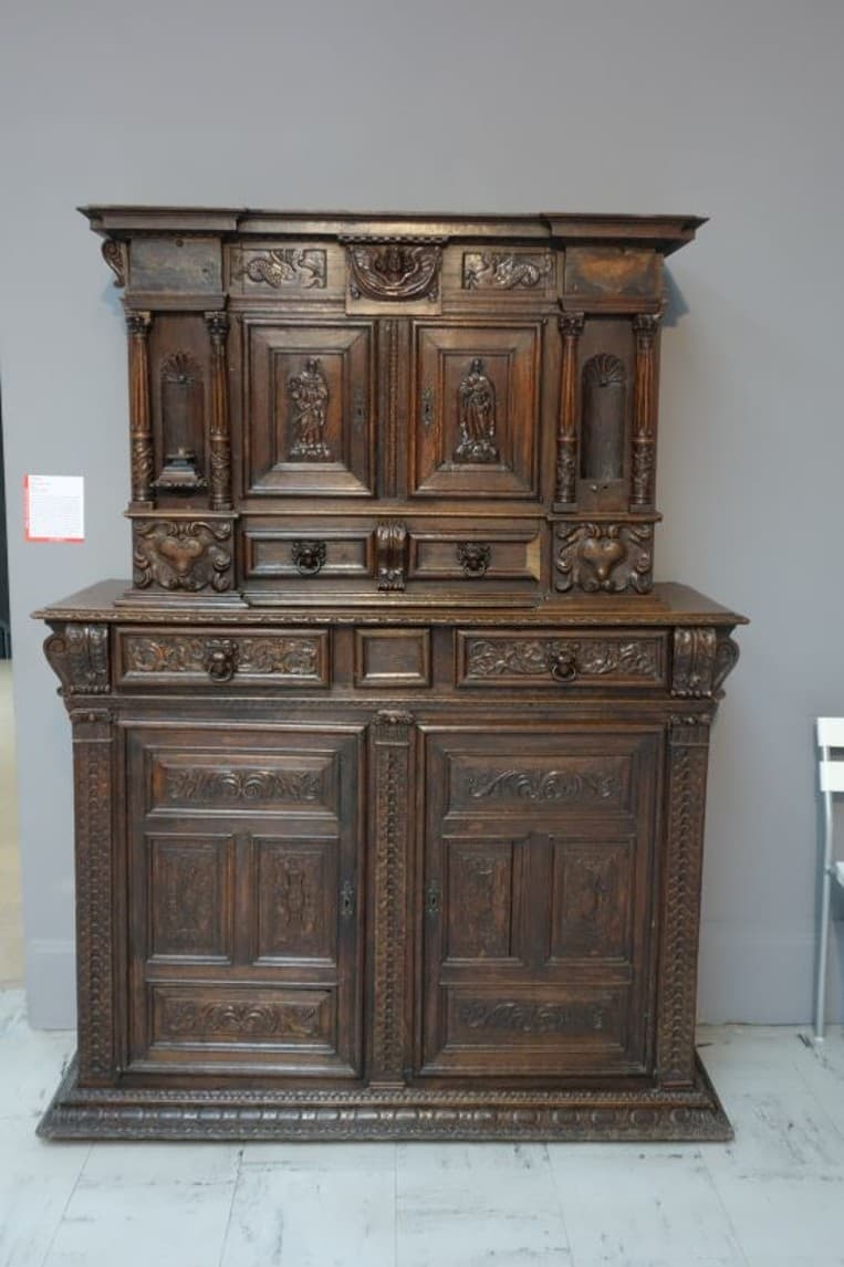 A tall, dark wooden cabinet with intricate carvings. The cabinet features a tiered design with multiple drawers and doors. The upper section has decorative columns and figural carvings, while the lower section has paneled doors with detailed relief work. The base and top edges are adorned with decorative molding. The overall impression is of ornate craftsmanship. — 3D model