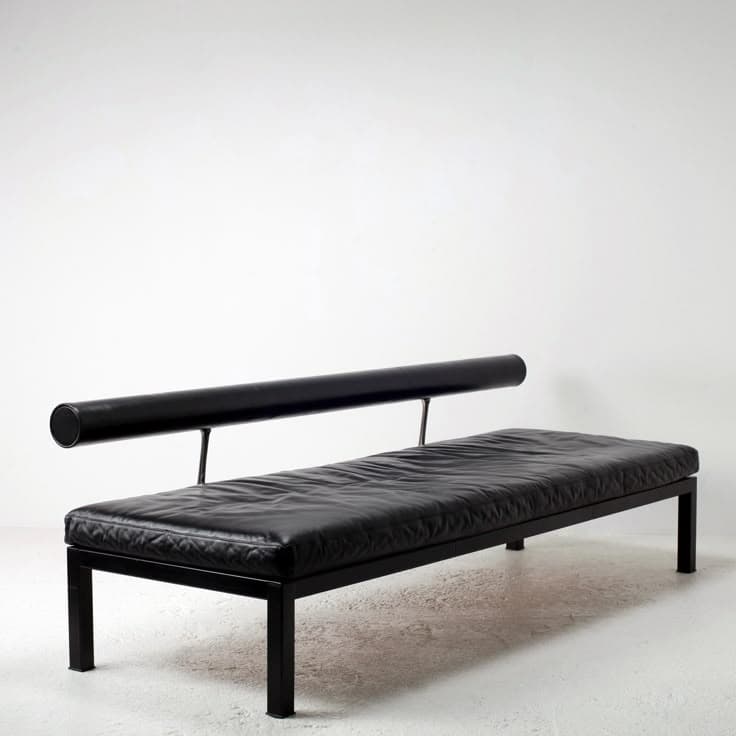A contemporary bench with a minimalist design. It features a rectangular upholstered seat and a cylindrical armrest or back support that extends horizontally along one side, supported by two thin vertical metal posts. The frame and legs are also made of metal. — 3D model
