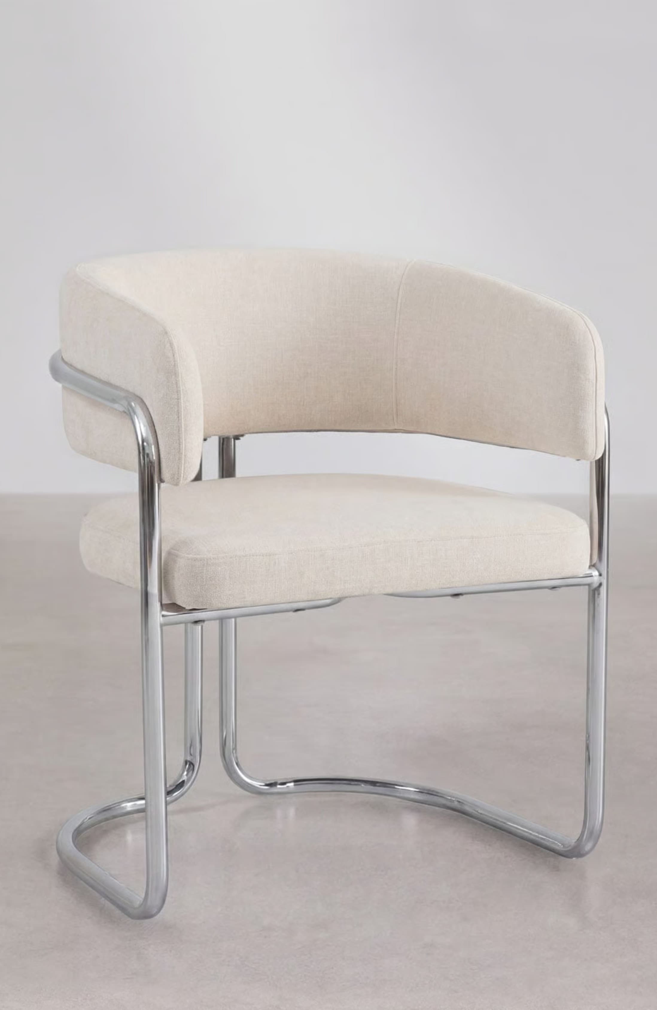 A modern armchair featuring a tubular chrome frame and upholstered in a light, neutral fabric. The seat and backrest are well-cushioned, and the back curves around to form integrated armrests. A polished chrome bar accentuates the exterior of the armrests, connecting to the continuous loop base. — 3D model