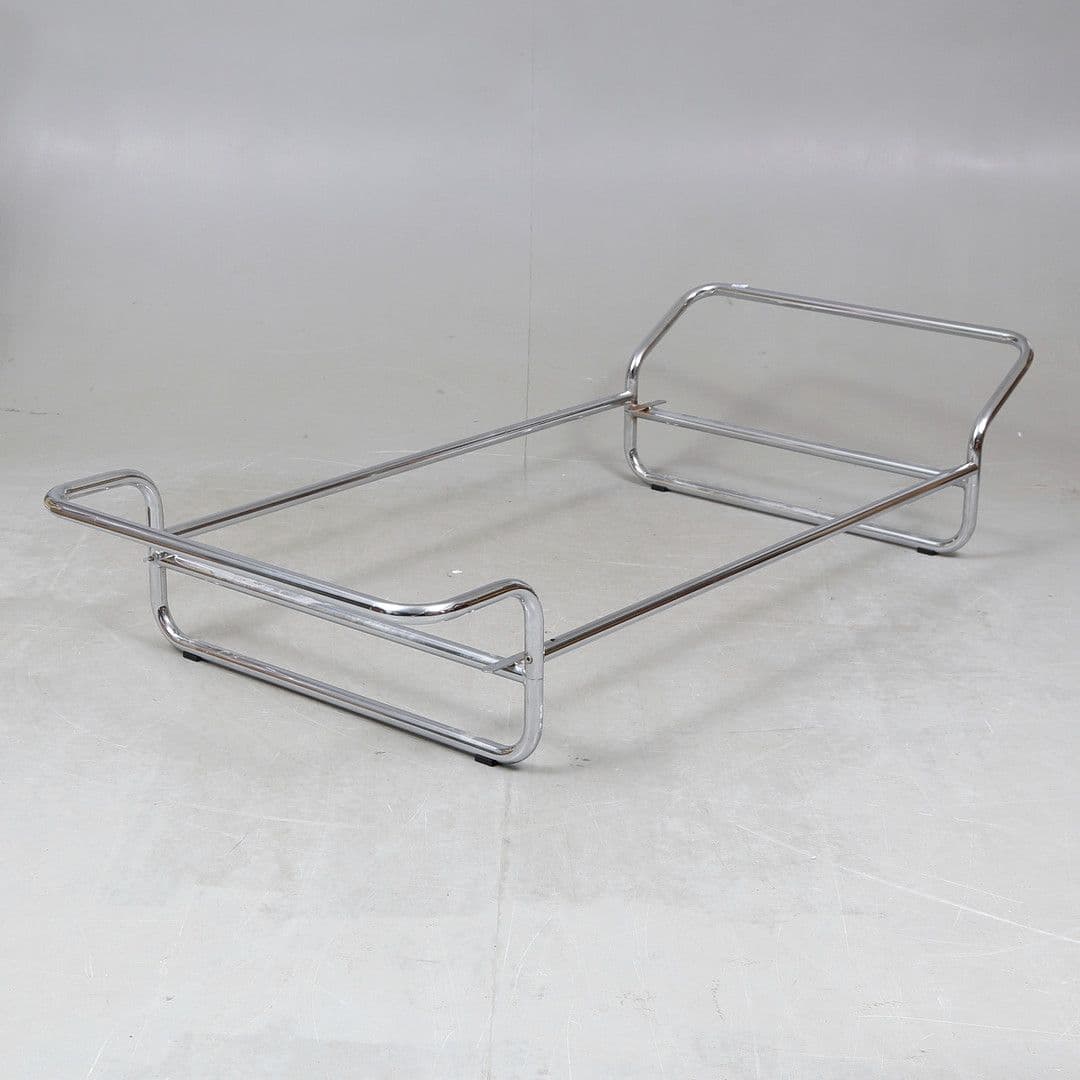 A minimalist bed frame constructed from polished chrome-plated tubular metal. It features a rectangular base with slightly raised ends, forming a headboard and footboard structure. The design is open and airy, with curved elements at the corners and a simple longitudinal support system. The frame rests on short, black cylindrical feet. — 3D model