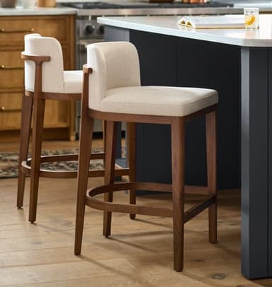 A modern bar stool featuring a dark-stained solid wood frame and light cream-colored fabric upholstery on both the seat and the gently curved backrest. The stool has four tapered legs with an integrated semi-circular footrest. — 3D model