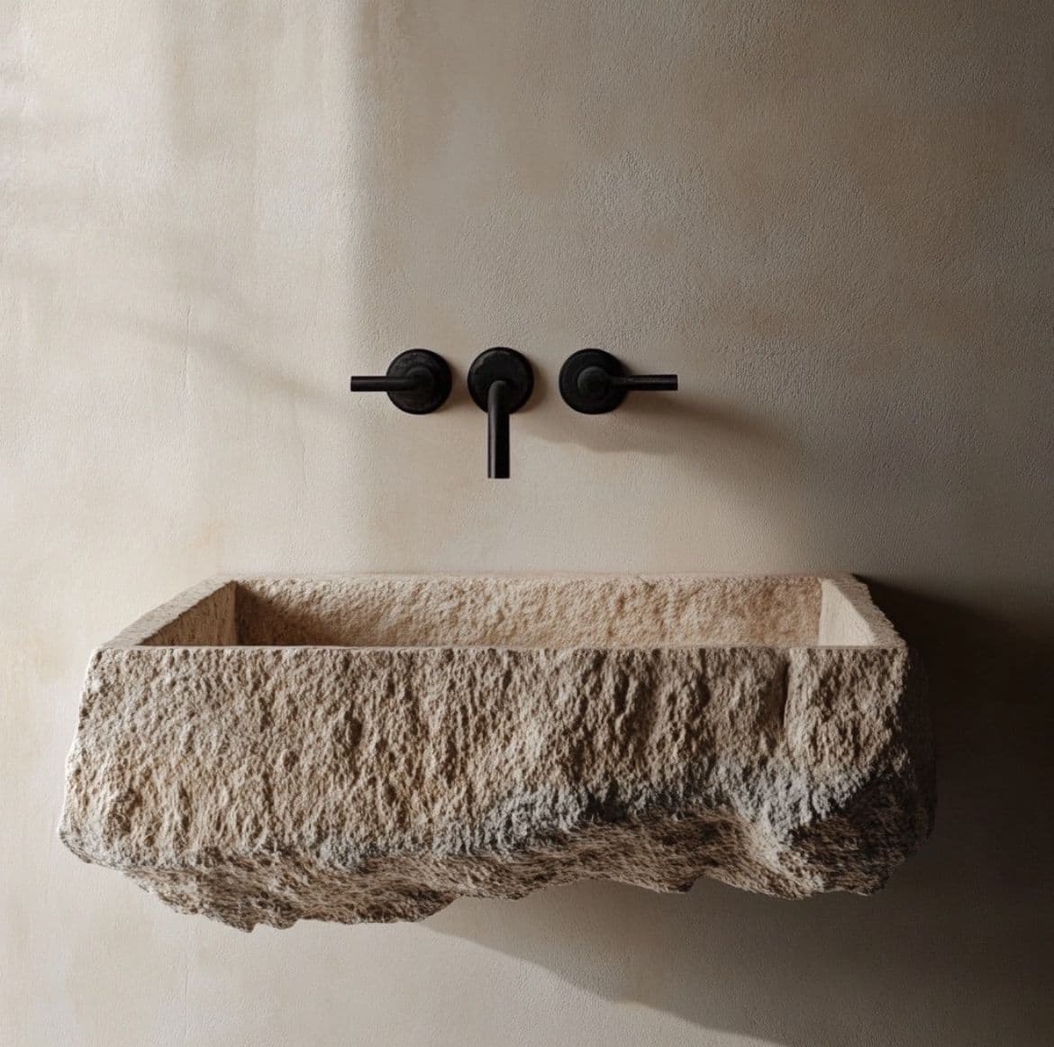 A rectangular stone vessel sink with a rough, natural exterior and a smooth interior. It features a black wall-mounted faucet with two handles and a spout, and a black drain cover within the basin. — 3D model