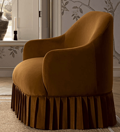 A plush armchair upholstered in a rich, matte brown velvet fabric. The armchair features a rounded back that seamlessly transitions into low, curved armrests. The base of the armchair is distinguished by a prominent, tailored pleated skirt that extends to the floor, adding a decorative and somewhat vintage aesthetic. The overall form is compact and inviting. — 3D model
