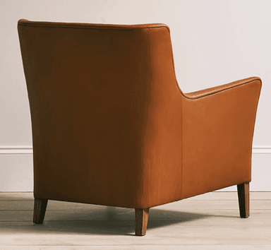 A contemporary armchair upholstered in cognac-colored leather, featuring a broad seat, low-profile rolled armrests, and a gently angled backrest. The chair is supported by four tapered wooden legs, visible beneath its substantial body. Visible stitching defines the edges and seams of the upholstery, contributing to its tailored appearance. — 3D model