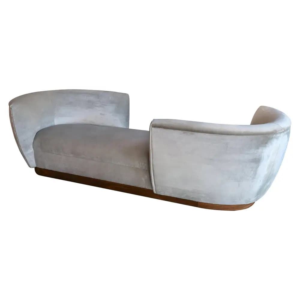 An elegant Art Deco style chaise lounge, featuring a streamlined, elongated form upholstered in plush light grey velvet, set upon a dark wooden base. The lounge is characterized by its inward-curving, raised ends that serve as both armrests and backrests, creating a symmetrical, cocoon-like silhouette. — 3D model