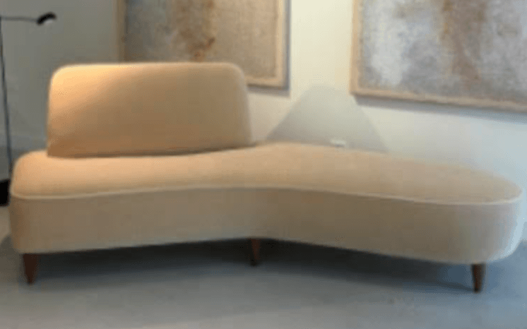 A modern asymmetrical chaise lounge upholstered in a light beige fabric with contrasting white piping, supported by tapered wooden legs. — 3D model