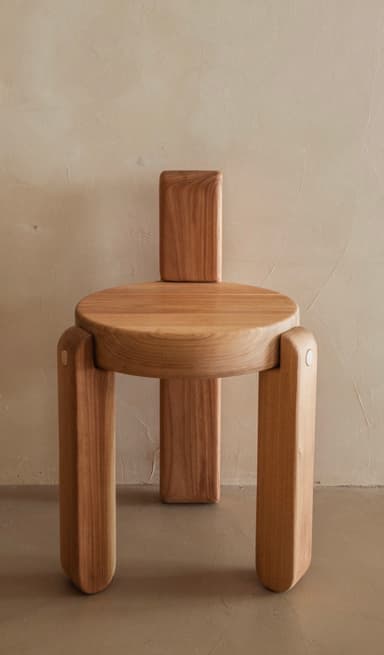 A minimalist, light-toned wood chair featuring a circular seat and a short, rectangular backrest. The design is characterized by thick, rounded elements for the legs and back, giving it a sculptural and contemporary appearance with visible wood grain. — 3D model