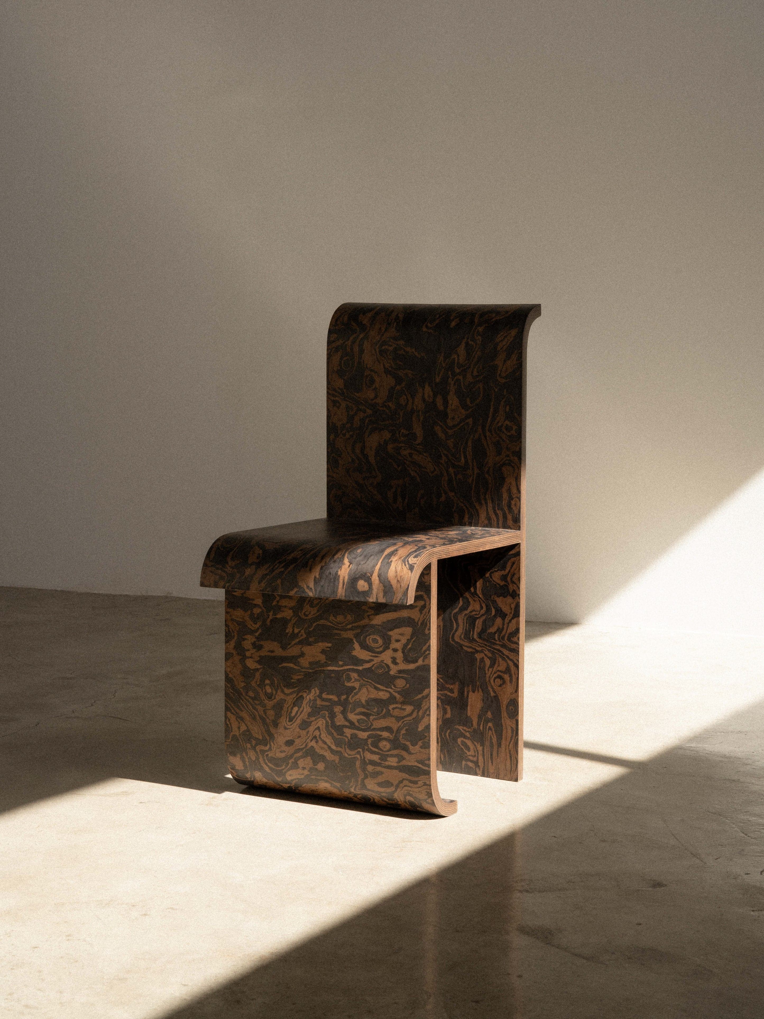 A minimalist, armless side chair constructed from a single piece of bent plywood, featuring a distinctive dark brown burl wood finish with swirling lighter brown patterns. The chair exhibits smooth, continuous curves that form the backrest, seat, and base, creating a sleek, sculptural silhouette. Its design emphasizes form and material. — 3D model