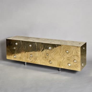 A long, rectangular, low-profile console cabinet or sideboard, finished in a brass-toned metal with intricate etched concentric circular patterns across its surfaces. Multiple small, clear, circular decorative inserts are embedded within the etched design. The unit stands on short, slender, silver-toned cylindrical legs. — 3D model