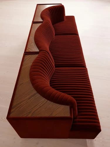 A contemporary, modular sofa with a striking, sinuous design. The sofa features a dark red, channel-tufted upholstery that flows in a wave-like pattern, contrasting with a base that appears to be made of wood with a visible grain. The structure is elongated and appears to be designed for multiple seating positions. — 3D model