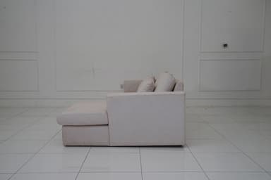 A sectional sofa with a chaise lounge extension, upholstered in a light-colored fabric with a subtle pattern. The sofa features three seat cushions and a chaise on the right side. It includes four matching throw pillows. The sofa has a modern, minimalist design with clean lines and low-profile arms and back. — 3D model