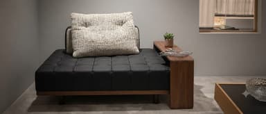 A contemporary low-profile daybed with black tufted leather upholstery, a minimalist wooden frame, and an integrated side table. A light-colored textured pillow is placed on the daybed, and a small potted plant and magazine rest on the wooden side table. — 3D model