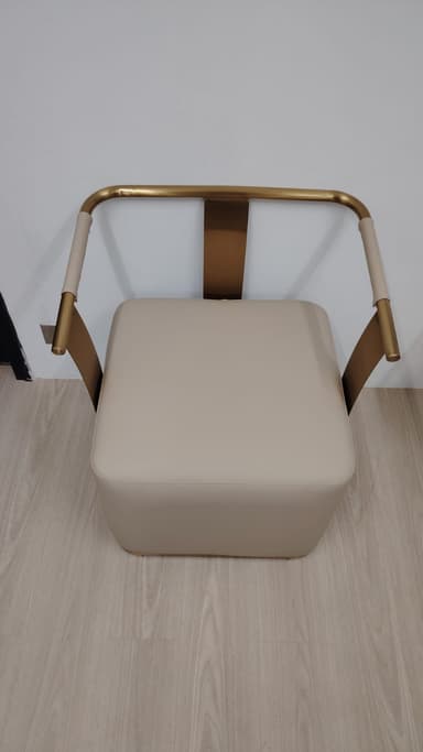 A modern armchair featuring a square, thick cushioned seat upholstered in a light beige material, supported by a minimalist brass-toned tubular metal frame that seamlessly forms the backrest and armrests. The design is sleek and contemporary. — 3D model