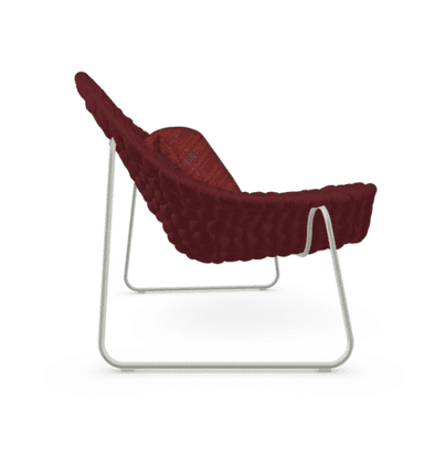 A modern lounge chair featuring a wide, scooped seating surface crafted from a deep maroon knitted or woven cord material, giving it a textured, sculptural appearance. The chair is supported by minimalist, light-colored metal wire frame legs. It includes a rectangular lumbar pillow in a complementary, slightly brighter red fabric. The overall design emphasizes comfort and a contemporary aesthetic. — 3D model