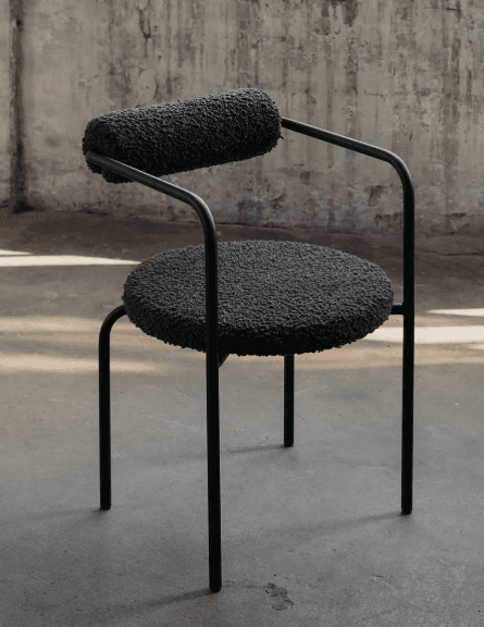 A modern chair with a rounded seat and a cylindrical backrest, both upholstered in a textured, dark gray fabric. The frame and legs are made of slender, black metal tubing. The armrests are formed by the continuous curve of the metal frame from the backrest to the front. — 3D model
