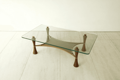 A modern coffee table featuring a rectangular clear glass top with softly rounded corners. The base consists of four sculptural, inverted-hourglass-shaped wooden legs, likely walnut, accented with brass rings. These legs support a brass frame that cradles a beige or taupe canvas sling, forming a secondary storage or display surface beneath the glass. The overall design suggests a blend of organic forms and metallic accents. — 3D model