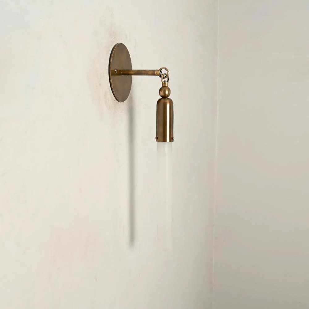 A modern wall sconce featuring a circular brass wall plate, an articulating arm, and a cylindrical brass housing holding a frosted glass rod-shaped shade. — 3D model