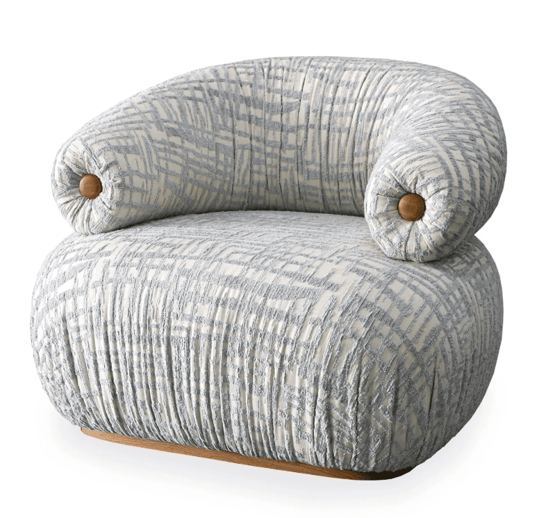 A contemporary armchair with a rounded, sculptural form. The chair features a high, curved back that flows into low-profile armrests. The upholstery is a textured fabric with an abstract geometric pattern in light blue and white. The armrests are accented with small, round wooden details. The chair is supported by a low, circular wooden base. — 3D model